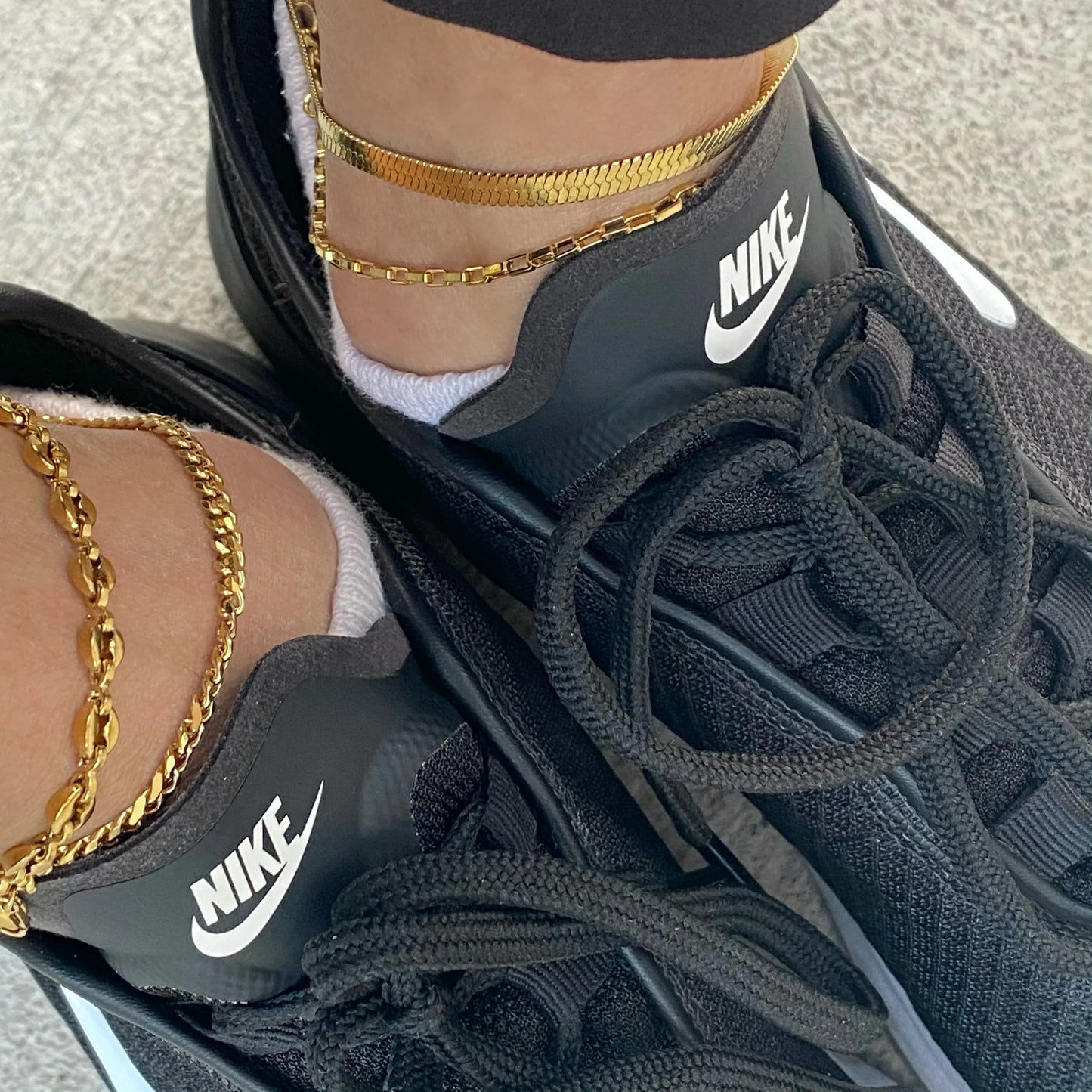 Close-up of feet wearing black Nike sneakers with white socks and gold chain anklets on gray ground