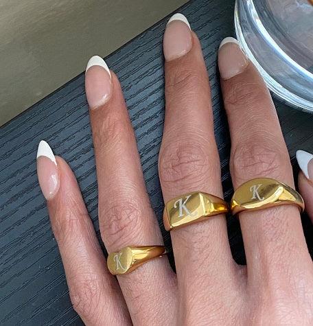 Hand with French manicure wearing three gold rings engraved with the letter K on black surface