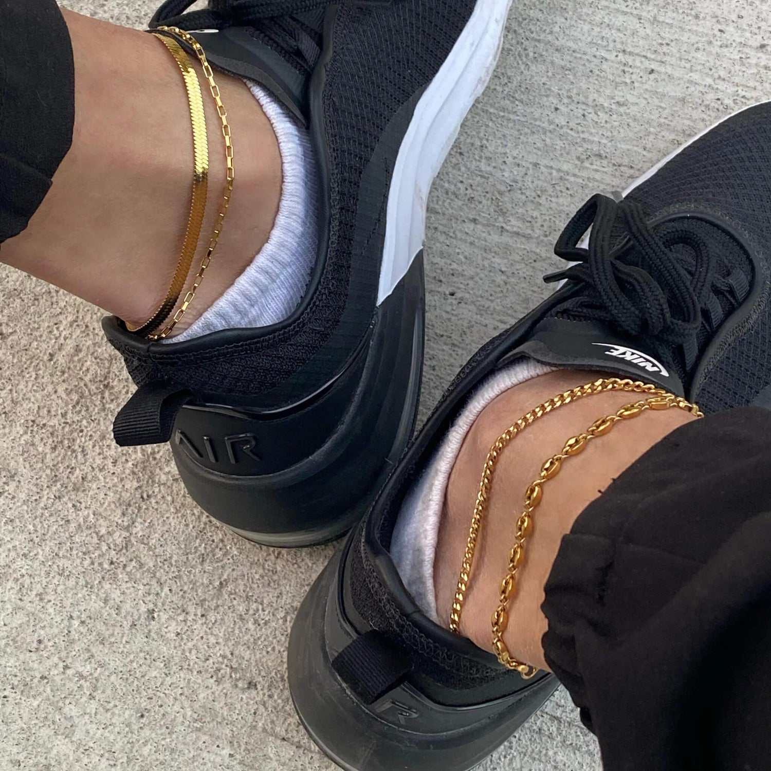 Person wearing black Nike Air sneakers and white socks with layered gold anklets on concrete floor