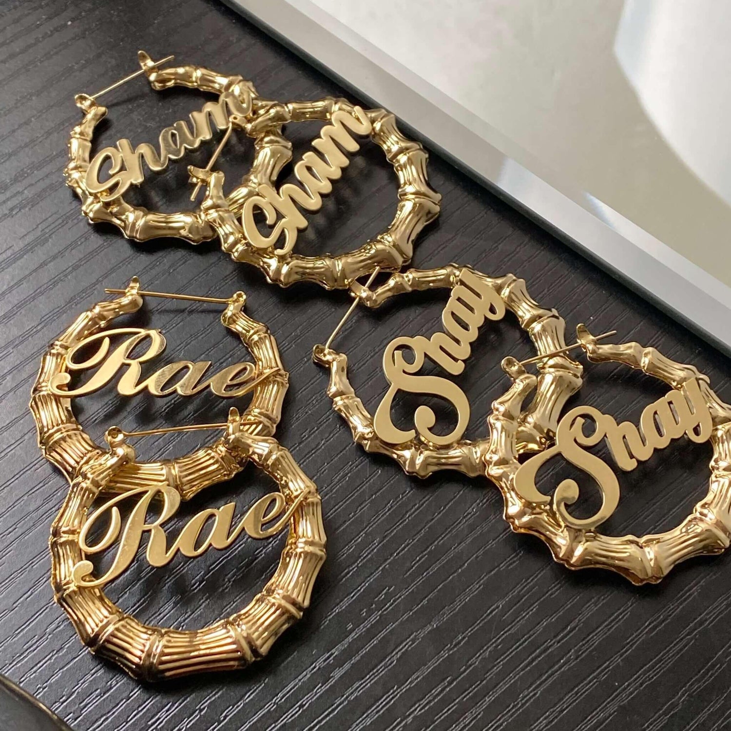 Gold personalized bamboo hoop earrings with names 'Rae' and 'Shay' on black surface