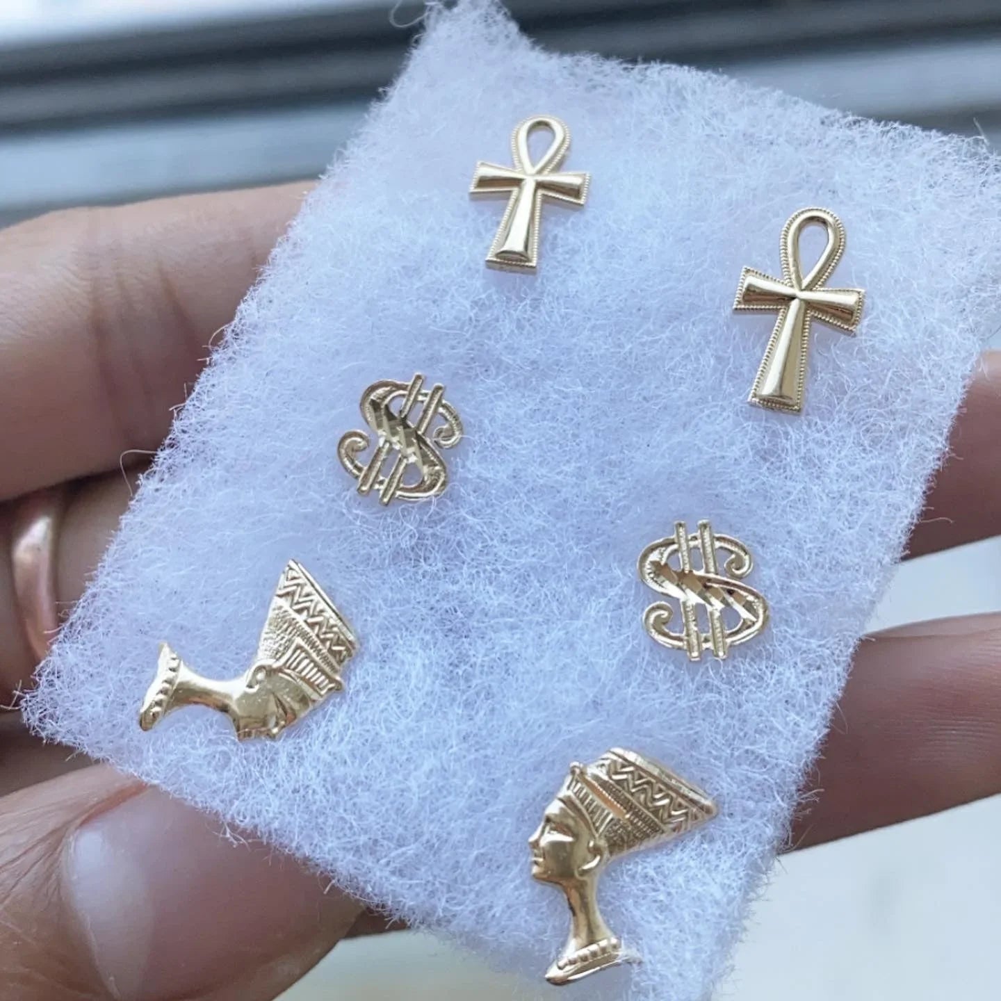 Gold stud earrings set with ankh, dollar sign, and Egyptian queen head designs on white foam